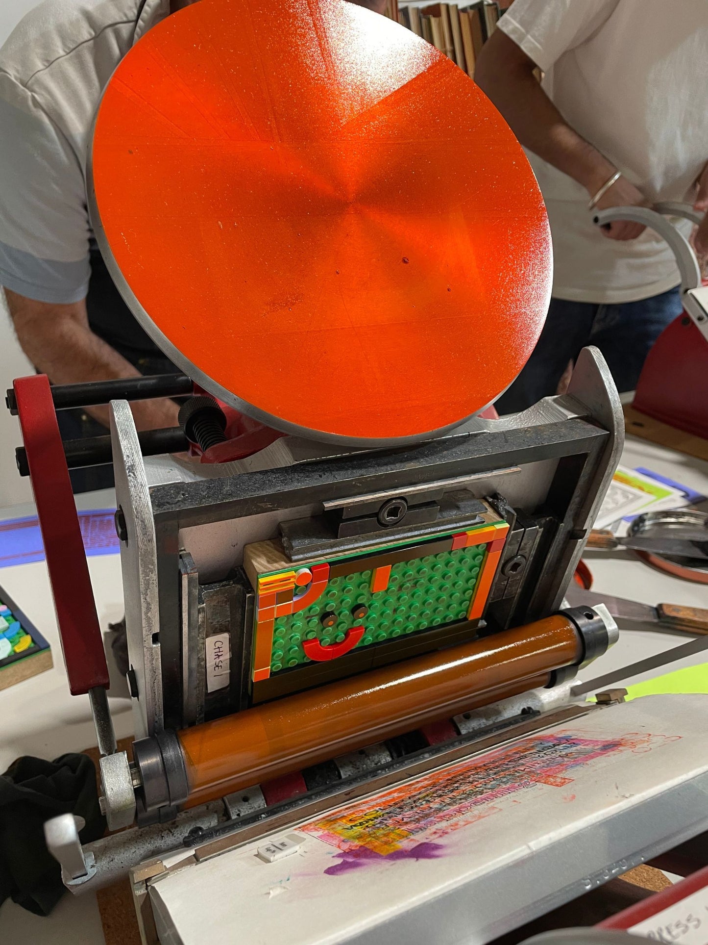 Close up of an Adana printing press with orange ink and a lego block in the press ready for printing.
