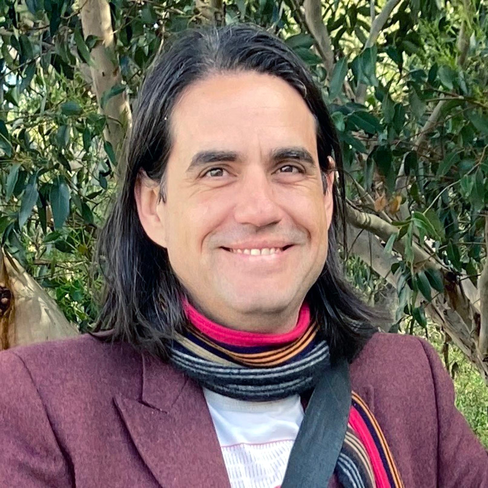 Chris Arnold, a man with long hair and a colorful scarf standing outdoors with trees in the background