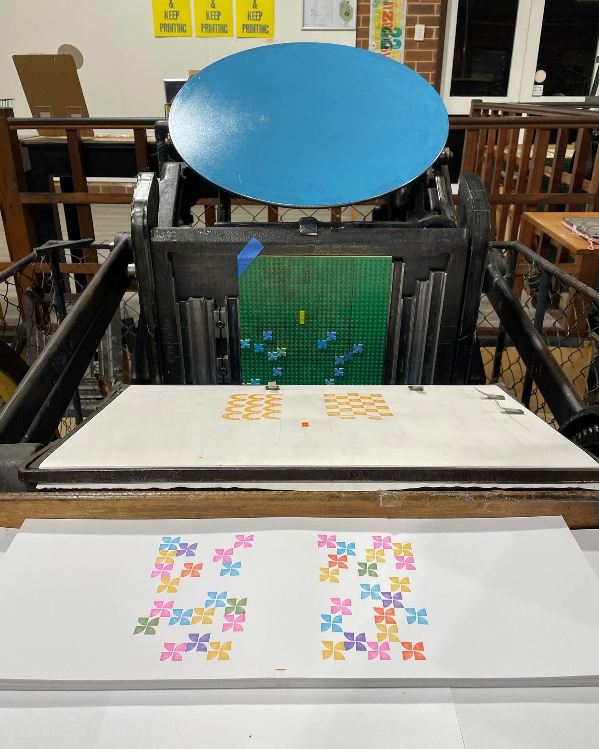 Printing press with blue inking disk and lego design locked in the press. Freshly printed prints on the feed table in front of the platen. 