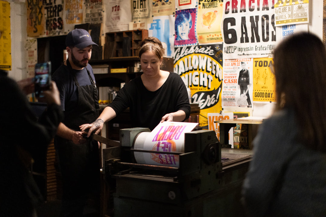 Person operating a printing press with posters on the wall in the background at the Moveable Type Studio