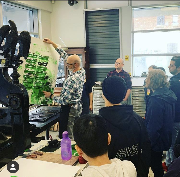 Phil Gambrill holding up a freshly printed print from a Columbian Press with a group of people in a workshop setting 