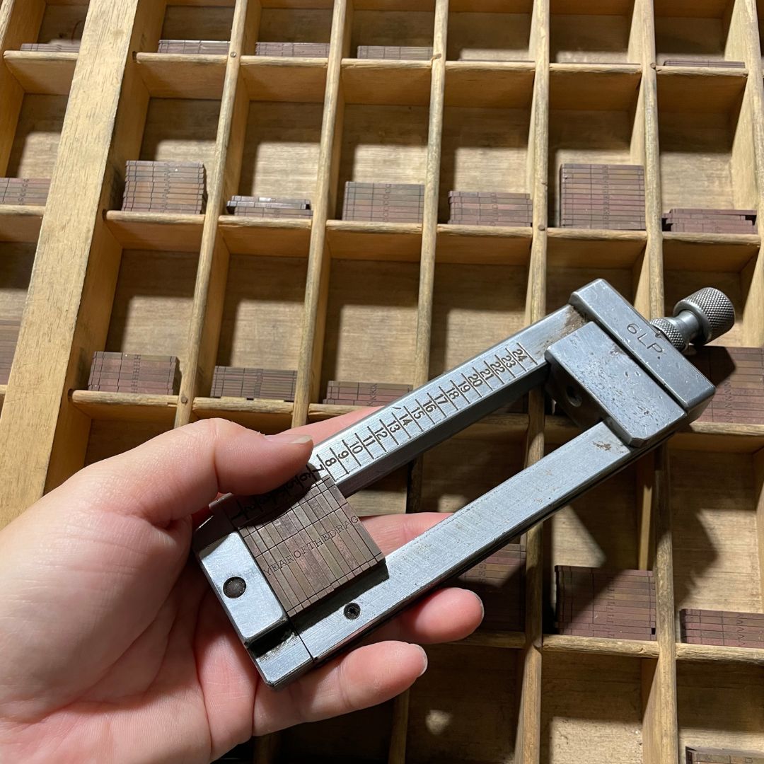 A hand holding the Ludlow composing stick in front of a case of Ludlow mats, in preparation to cast lines of metal type.