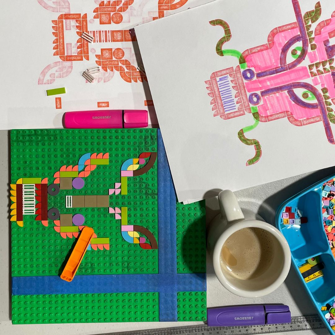 Bird’s eye view of some of the work in progress. There is a Lego base plate with Lego tiles positioned and creating a part silhouette of a dragon’s head. There are a couple of test prints surrounding the Lego base plate, along with a box of Lego tiles, highlighters and a cup of coffee. 