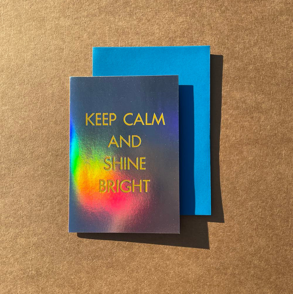 Greeting card with 'Keep Calm and Shine Bright' text on a rainbow holographic foil card, placed on a blue envelope against a brown surface.
