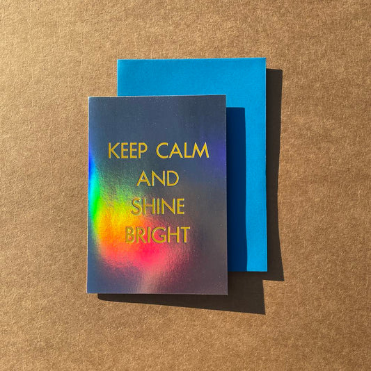Greeting card with 'Keep Calm and Shine Bright' text on a rainbow holographic foil card, placed on a blue envelope against a brown surface.