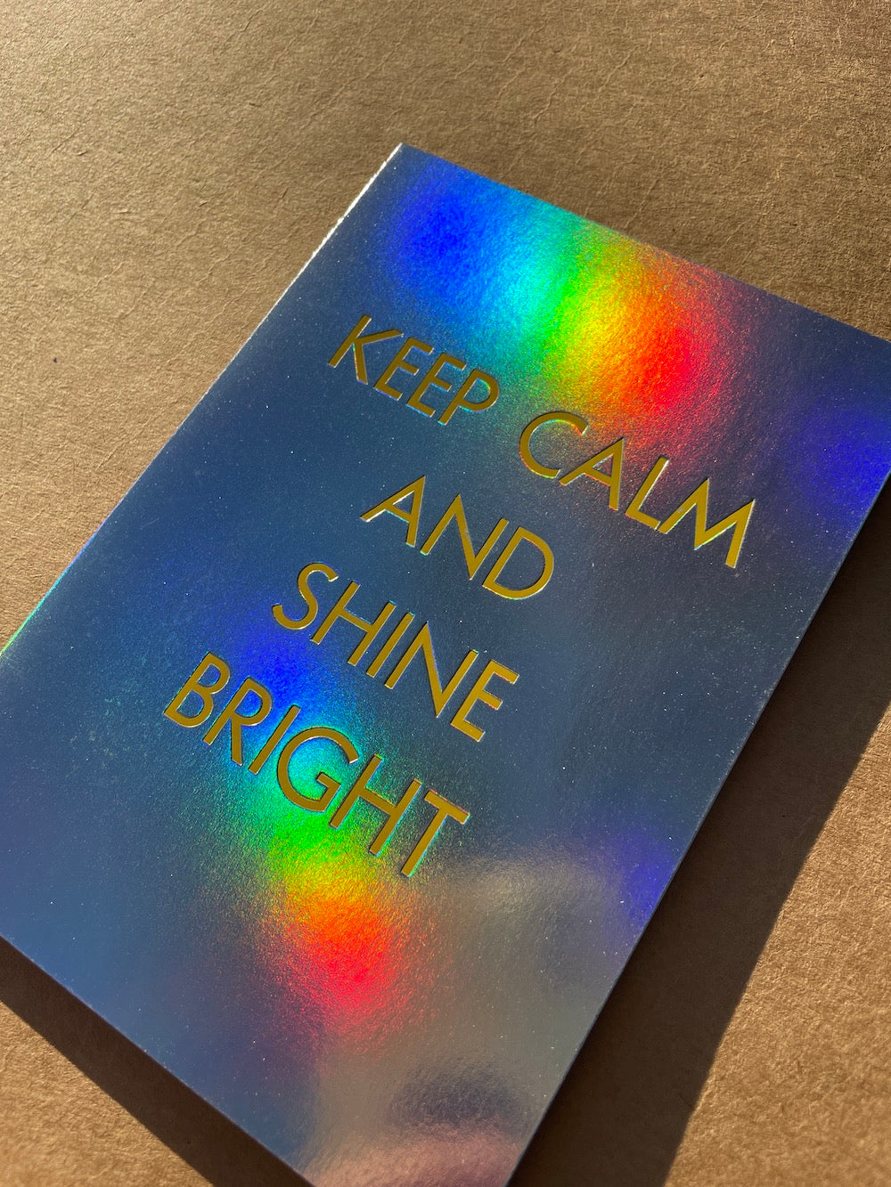 Close up of Holographic foil card with 'Keep Calm and Shine Bright' text on a brown background
