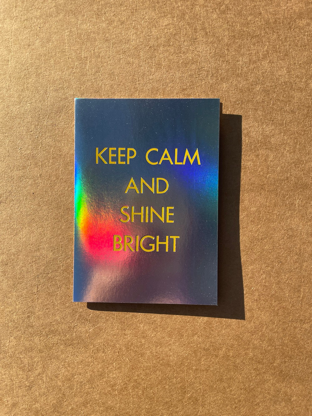 Rainbow holographic foil card with 'Keep Calm and Shine Bright' text on a textured brown background