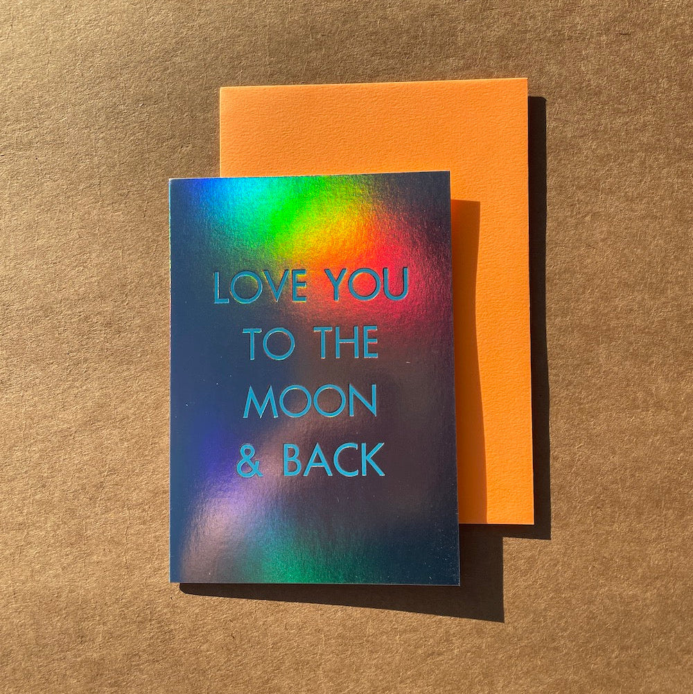 Greeting card with 'Love you to the moon & back' text on a rainbow holographic foil card, placed on an orange envelope against a brown textured background.