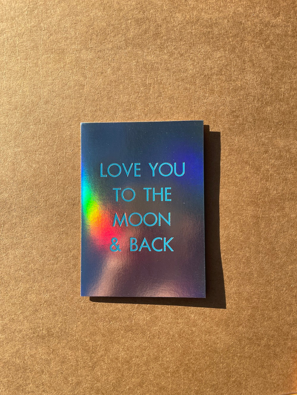 Rainbow holographic foil card with 'Love you to the moon & back' text on a textured brown background