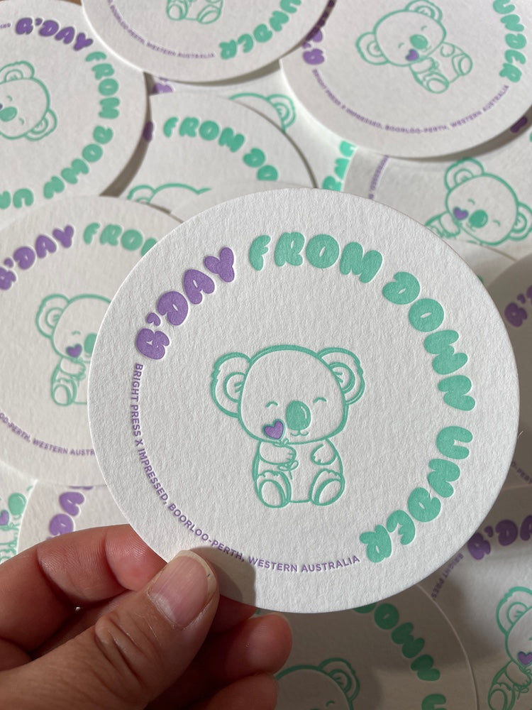 Circular coaster with a koala design and text held by a hand, surrounded by more coasters.
