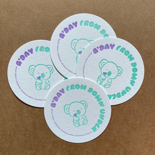 Four round coasters with a koala design and text on a brown background