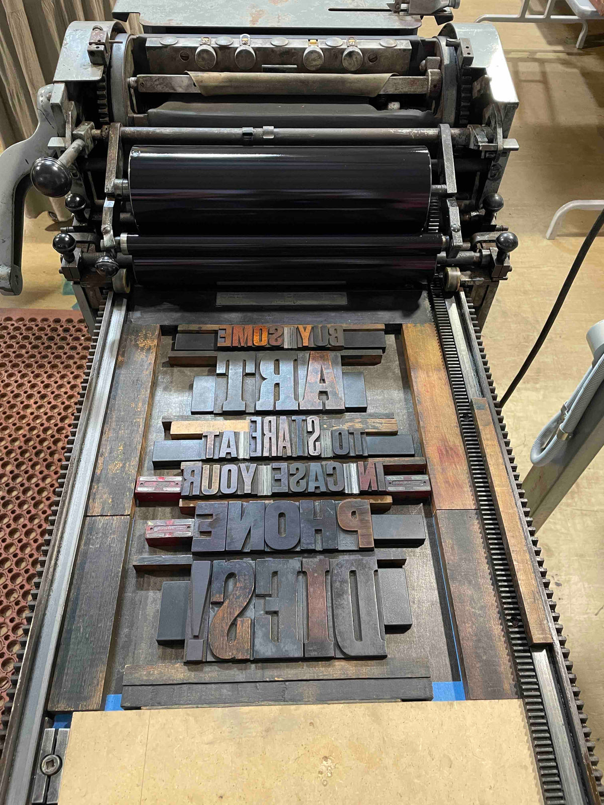 A bird's eye view of wood type on the press. Backward text reading "Buy some art to stare at in case your phone dies!"