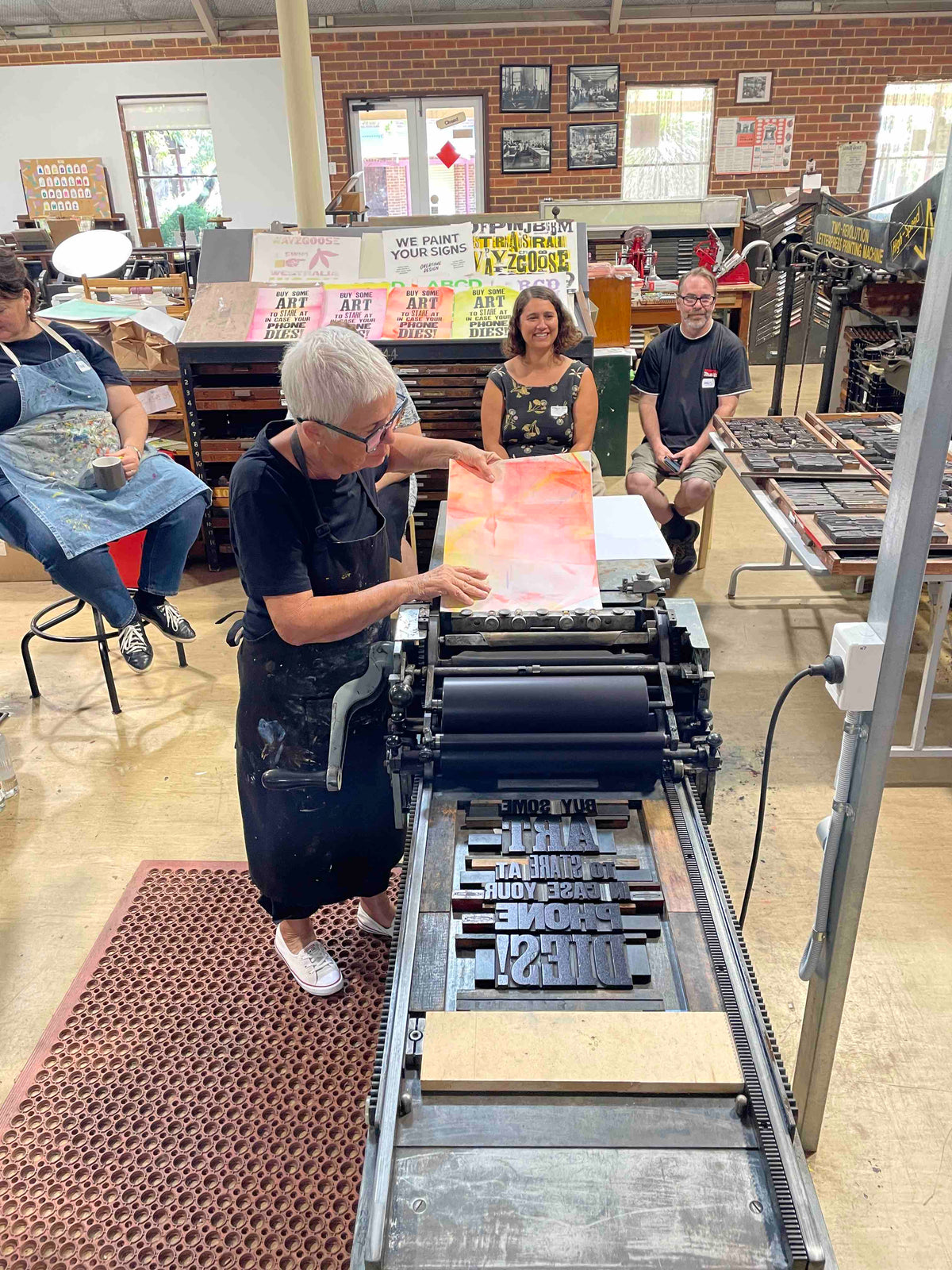 A participant feeding a sheet of red-orange card into the press in preparation to print their poster. Several participants are sitting around the press.