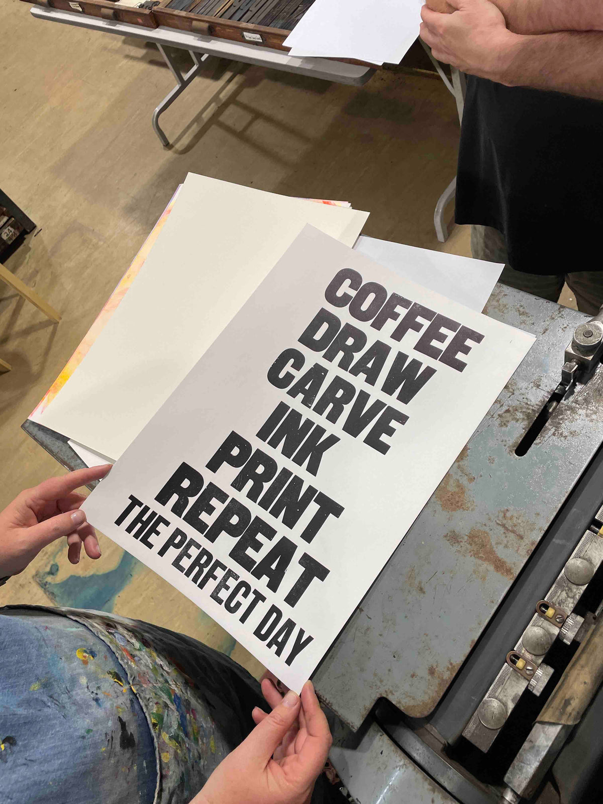 a bird's eye view of a freshly printed poster. Text reading "Coffee, Draw, Carve, Ink, Print, Repeat, The Perfect Day".
