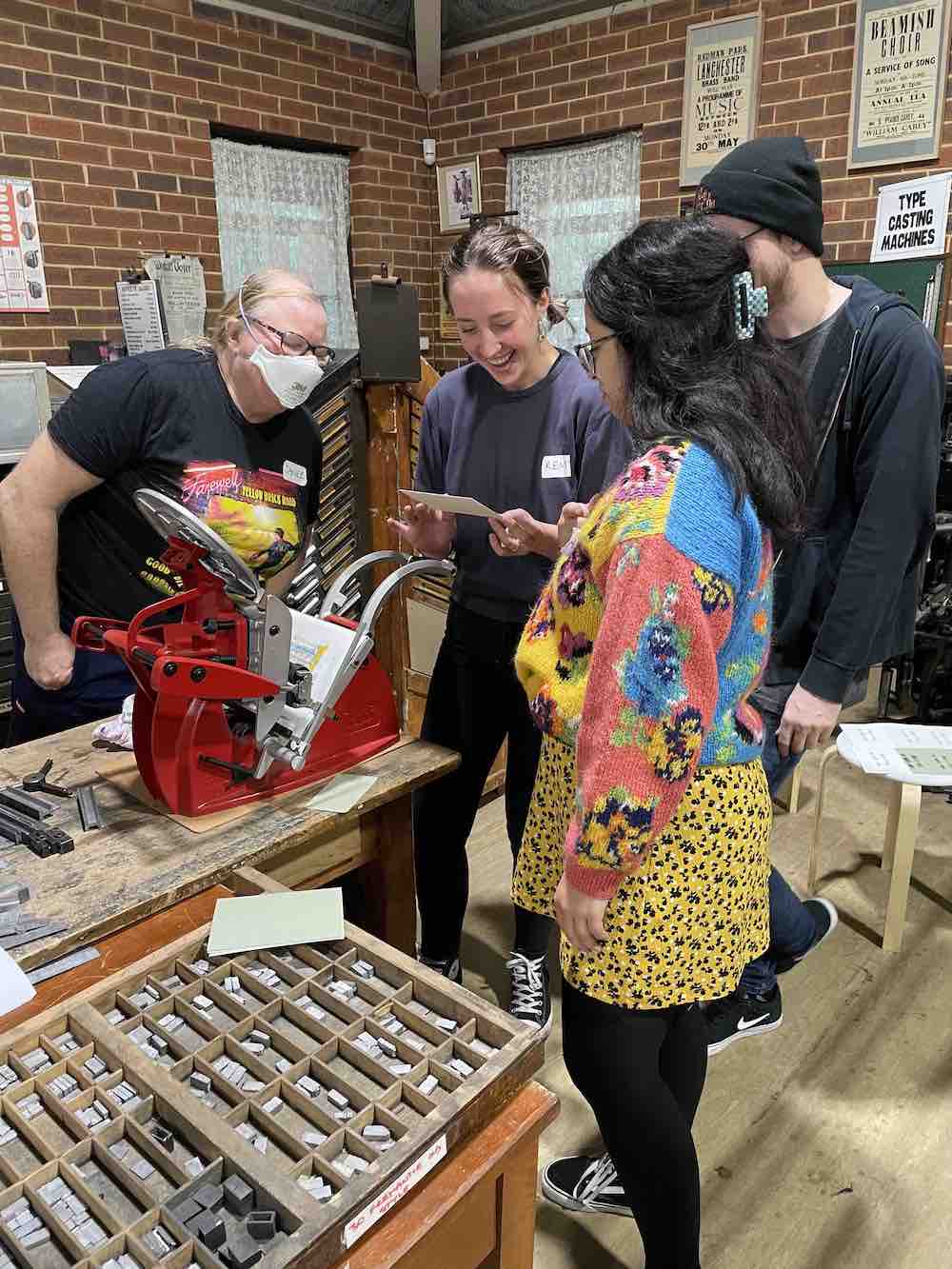 Four participants gathering around to check one of their freshly printed creations in front of an Adana press.