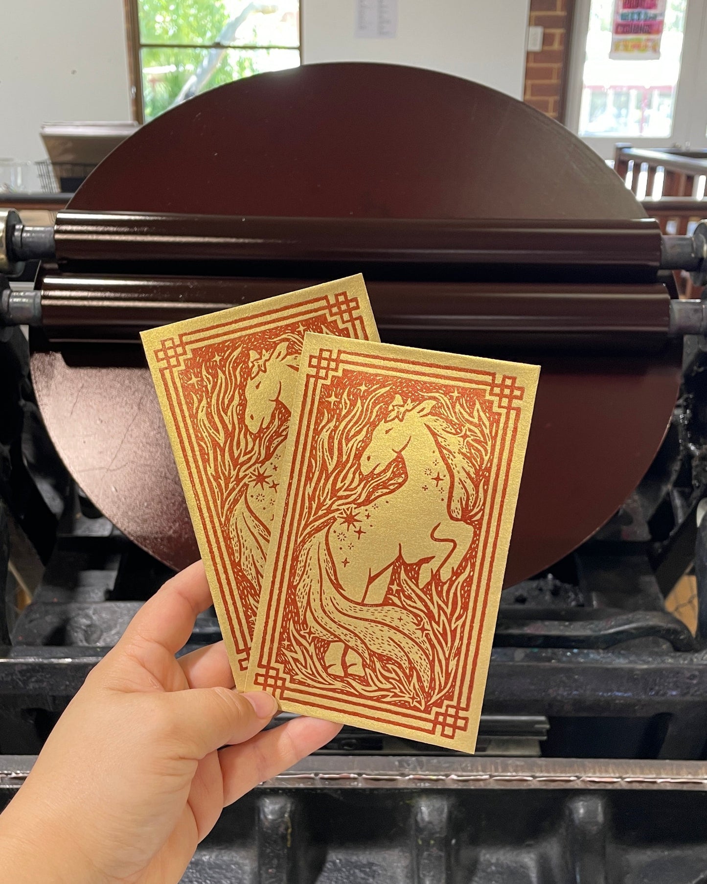 Hand holding two gold envelopes with a horse design in front of a printing press.