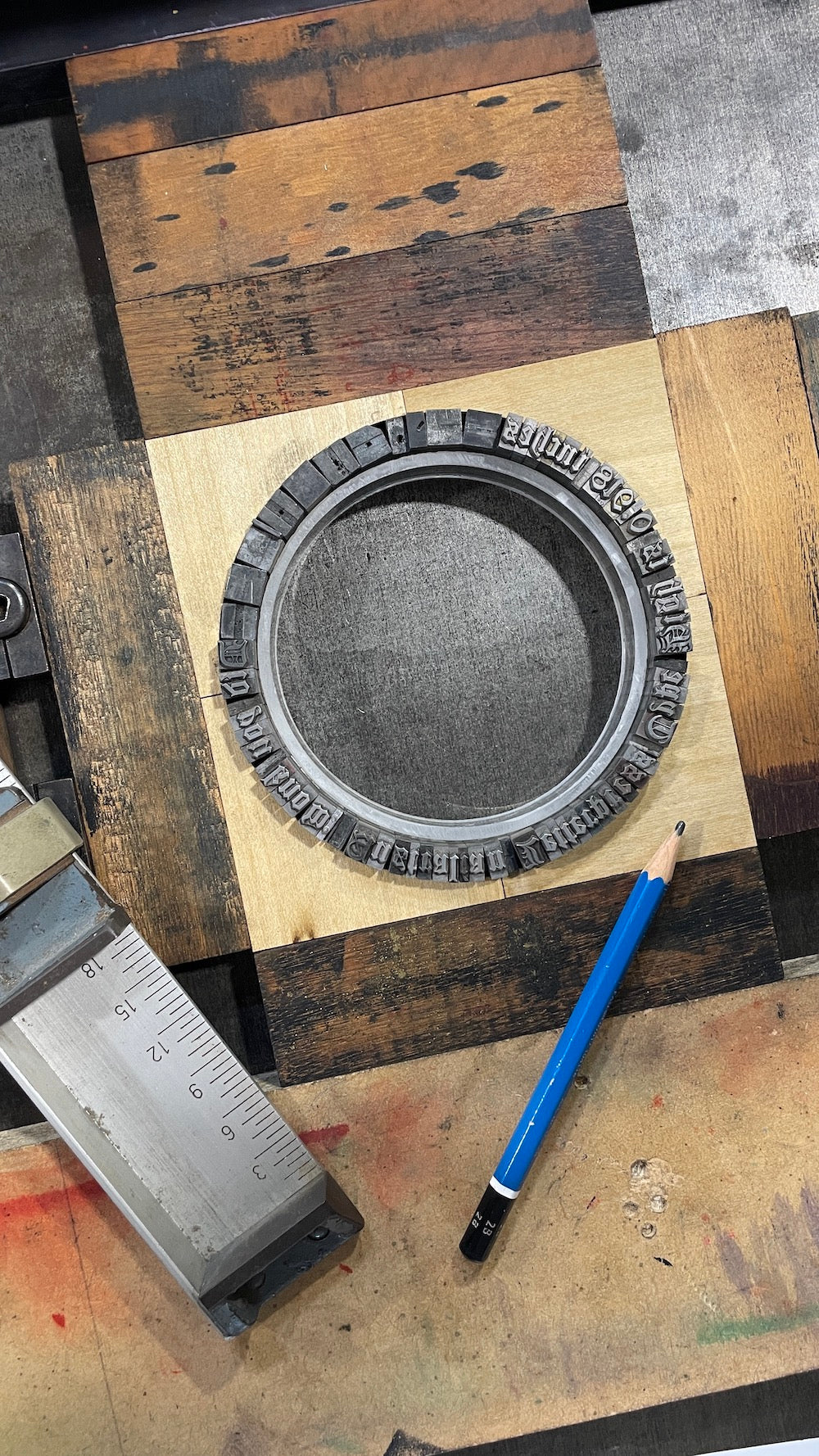 A circular forme with metal type, placed on the bed of a press with  wooden furniture alongside a blue pencil and a metal composing stick.