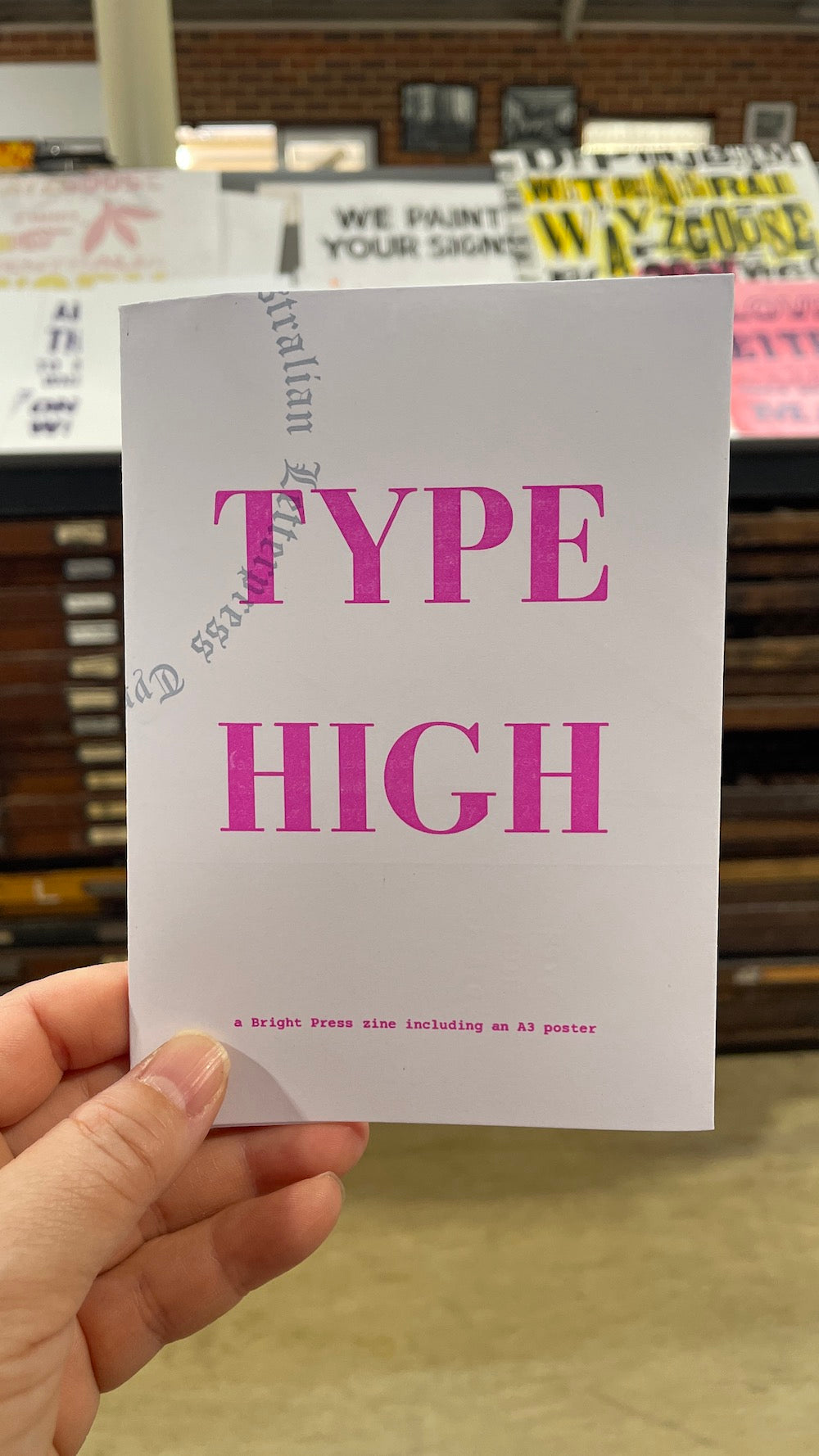 A hand holding a white zine with the words 'TYPE HIGH' in bold pink letters, with a blurred bookstore or shop interior in the background.
