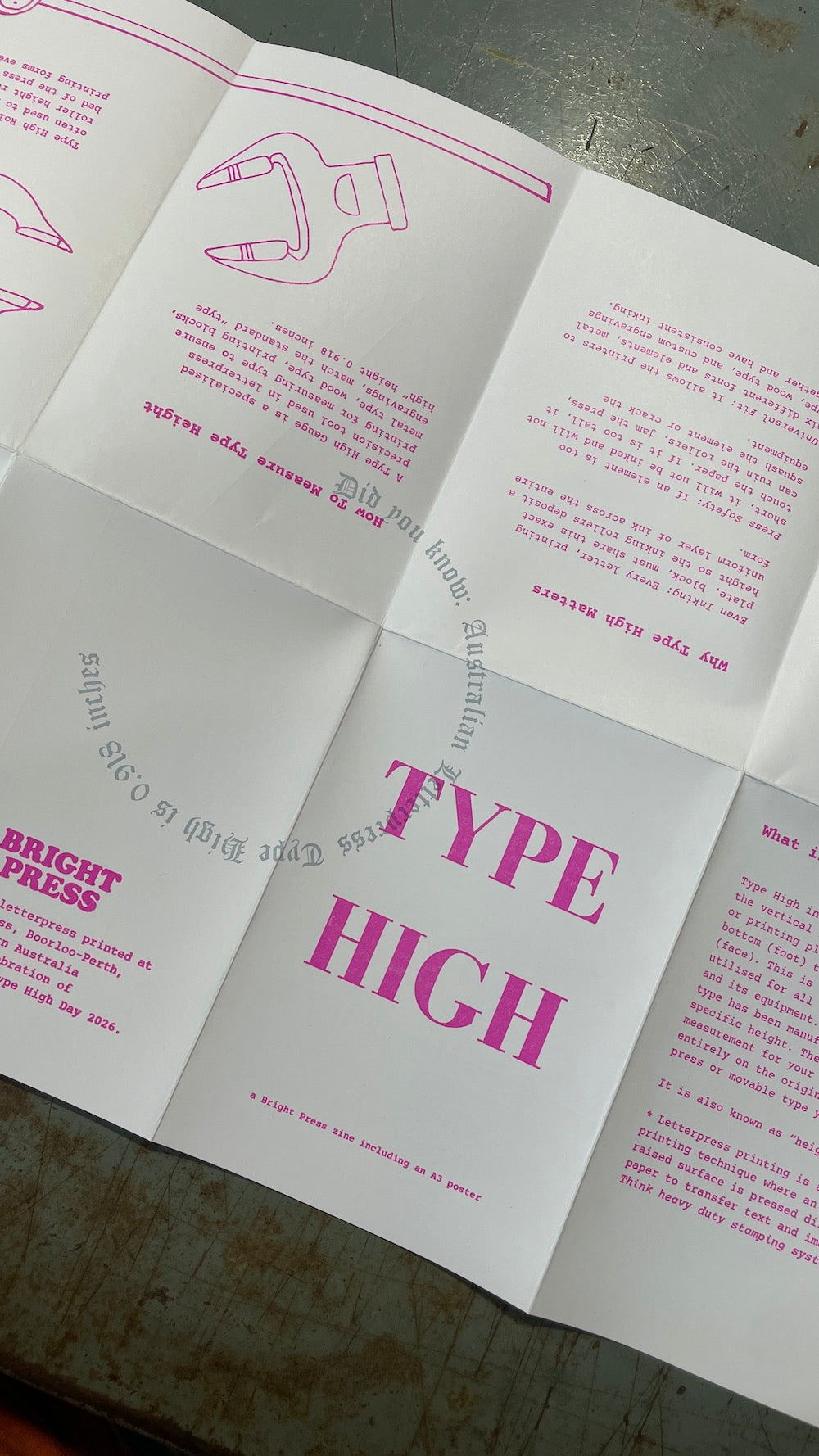 An open posterzine laid on a dark tabletop, showing white pages with pink text and line drawings. The center panel prominently reads “TYPE HIGH,” and other visible text includes “BRIGHT PRESS.