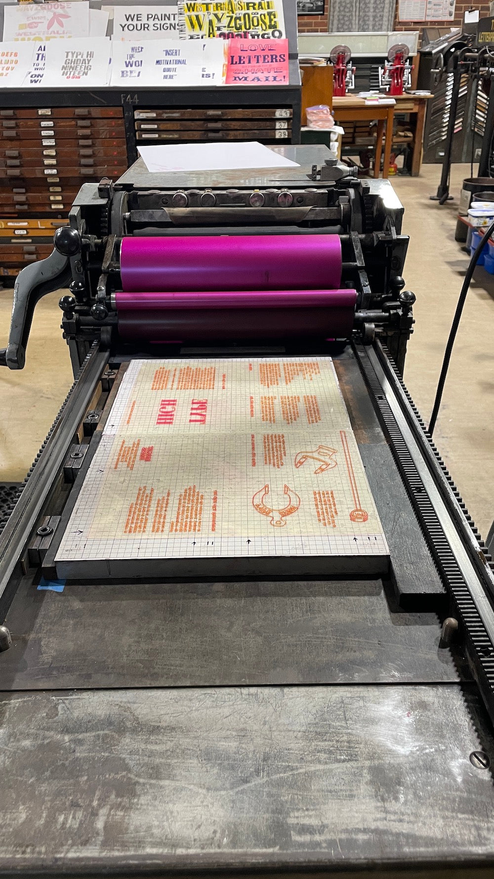 A large vintage-style printing press with a fluoro magenta ink on the rollers, with a mounting base on the press bed that has the polymer plate of the zine text inked up with the pink ink.