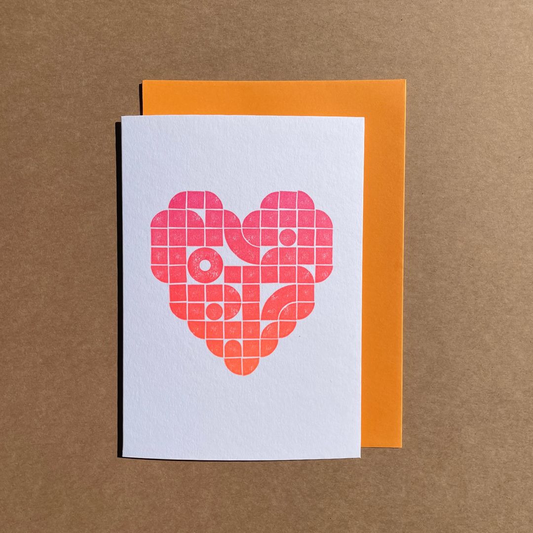 A love heart letterpress printed in the middle of the greeting card using lego tiles and in a gradient of multi-colours from pink to orange. The card is paired with an orange envelope.