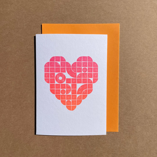 A love heart letterpress printed in the middle of the greeting card using lego tiles and in a gradient of multi-colours from pink to orange. The card is paired with an orange envelope.