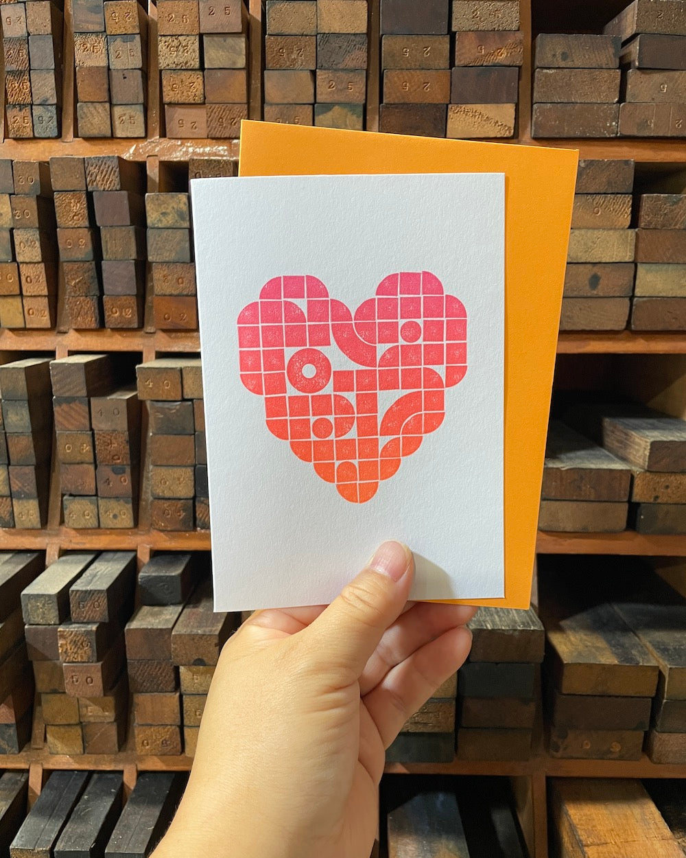 A hand is holding the card and envelope. In the background is a letterpress furniture rack