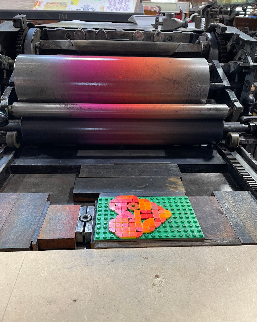 The heart design created with lego tiles is in the printing press and inked up ready for printing. The printing rollers have a gradient hue of pink, red and orange.