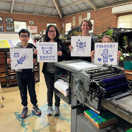 Four attendees (two young people along with their parents) holding up their final lego letterpress prints behind our Vandercook press.