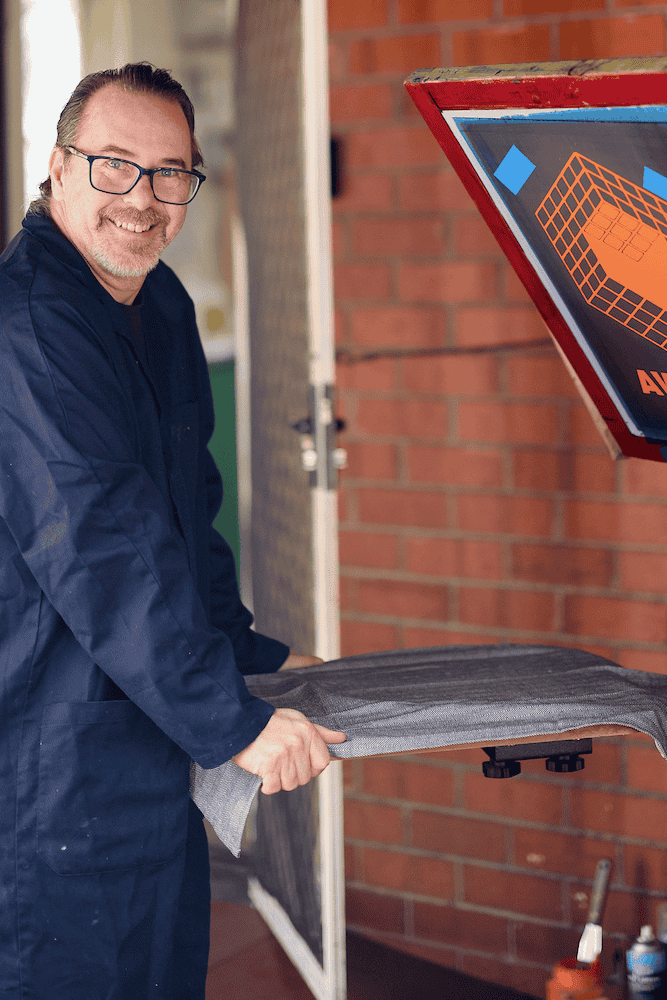 P. James Bryans in a blue jumpsuit holding a screenprinting platen next to a brick wall.