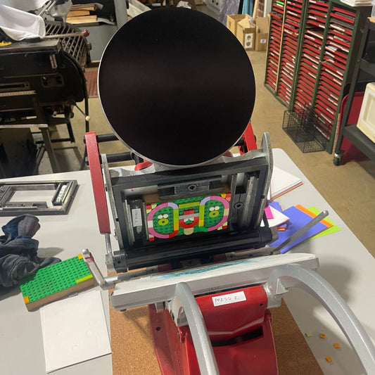 An Adana press with black ink and a lego block with a design freshly created by a young person in the chase and ready to be printed.
