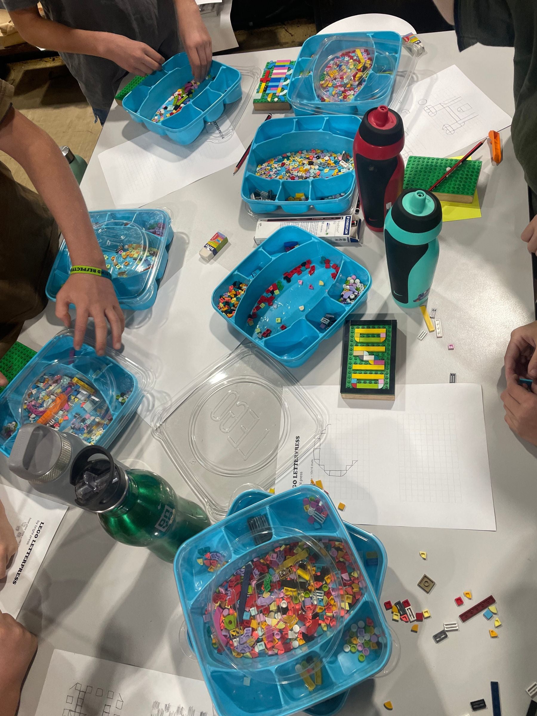Bird's eye view of a table full of boxes of lego tiles and papers with draft designs created by young people, and many pairs of hands getting stuck into the creating process with their lego blocks.
